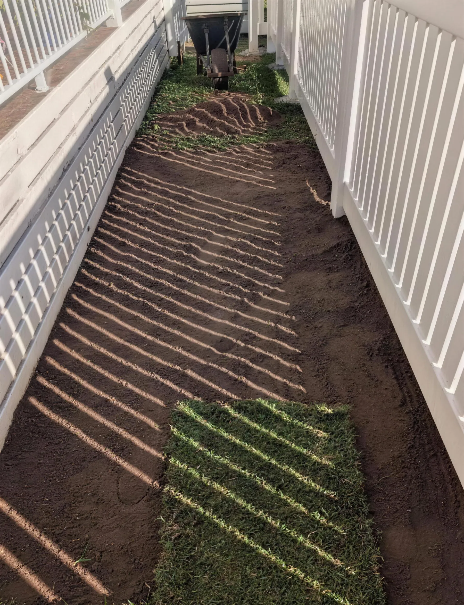 Fresh turf rolls being laid over prepared soil beside a white fence